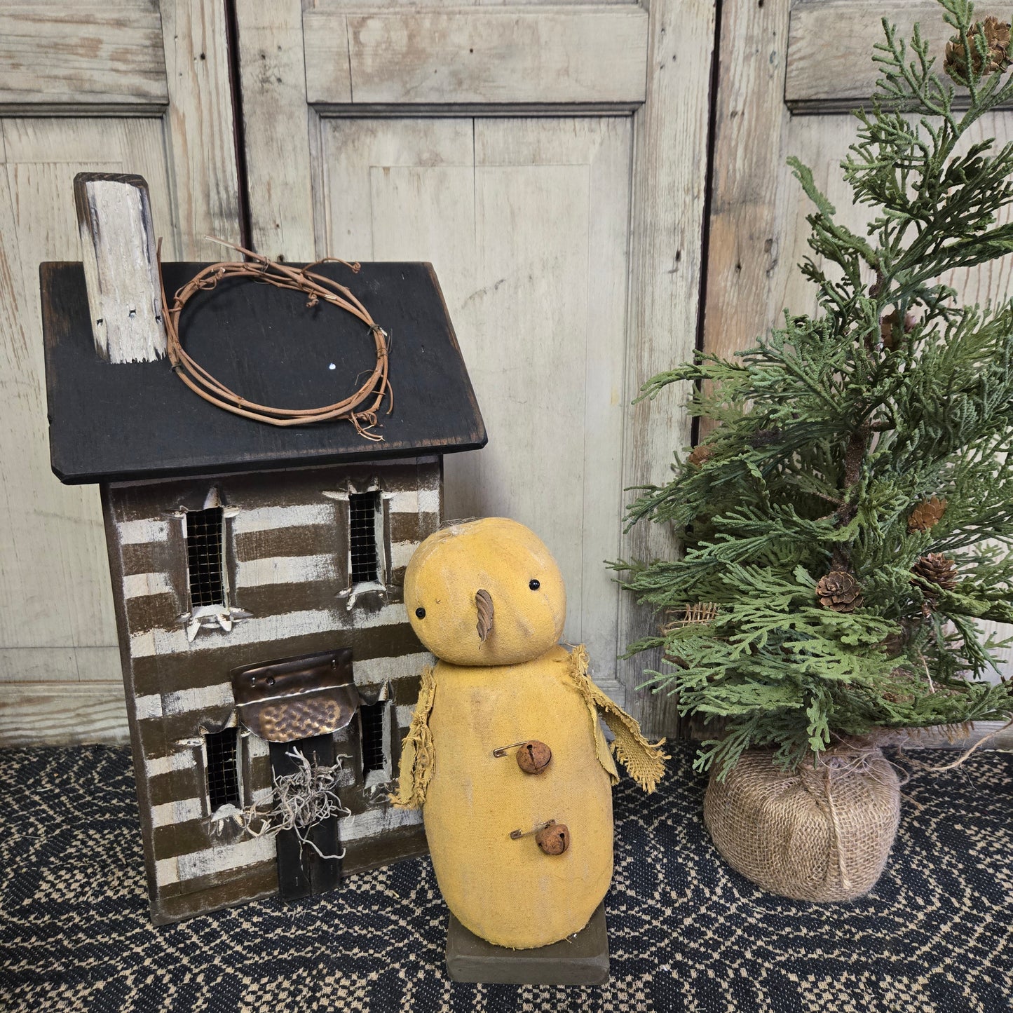 A handcrafted primitive chick with rusty bells figurine, displayed next to a small wooden house and a greenery decoration.