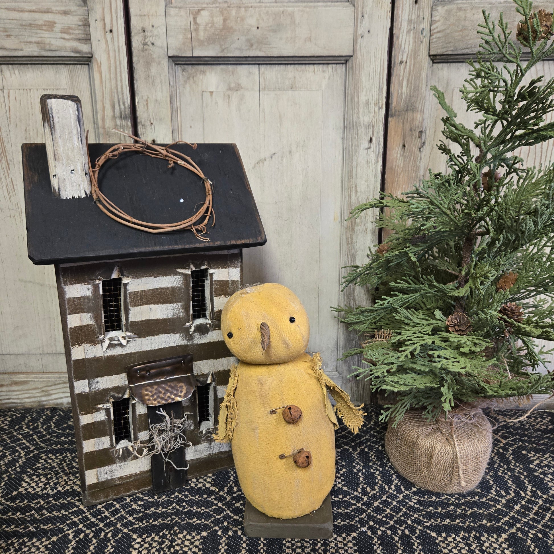 A handcrafted primitive chick with rusty bells figurine, displayed next to a small wooden house and a greenery decoration.