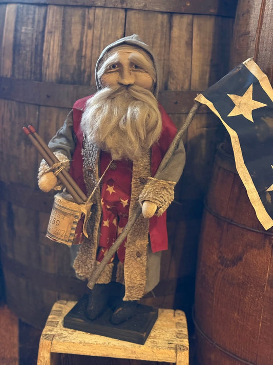 Statue of a bearded man with a hat and coat, holding a flag and walking stick, standing on a wooden block with barrels in the background.
