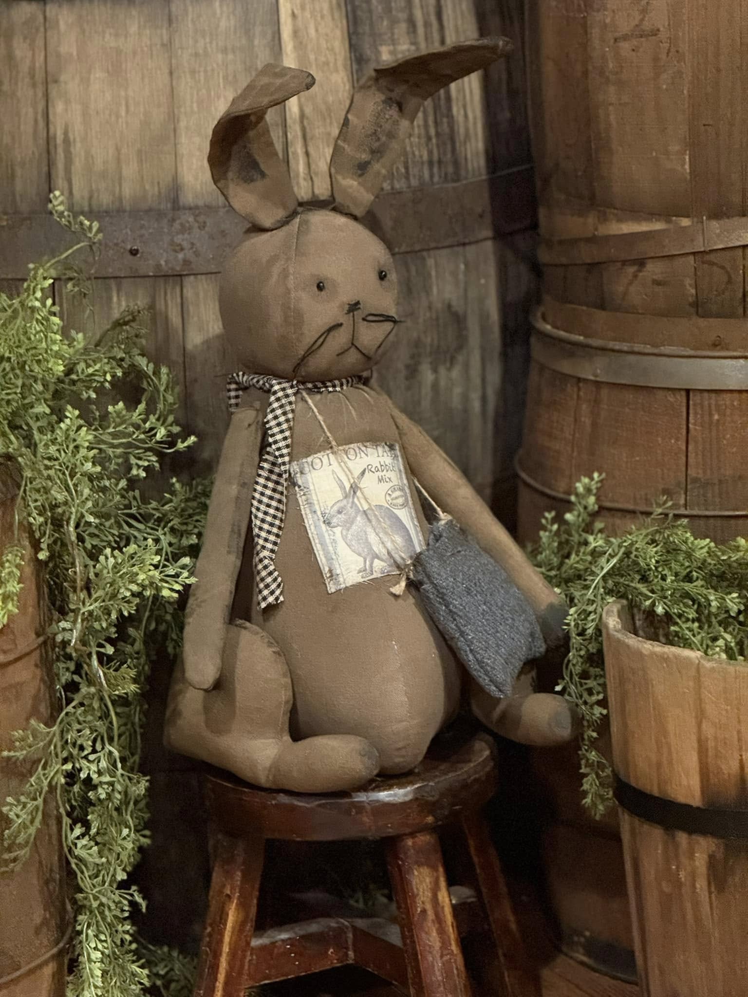 A handcrafted Primitive Cottontail Bunny doll with a brown body, beige details, and a checkered scarf, seated against a rustic background.