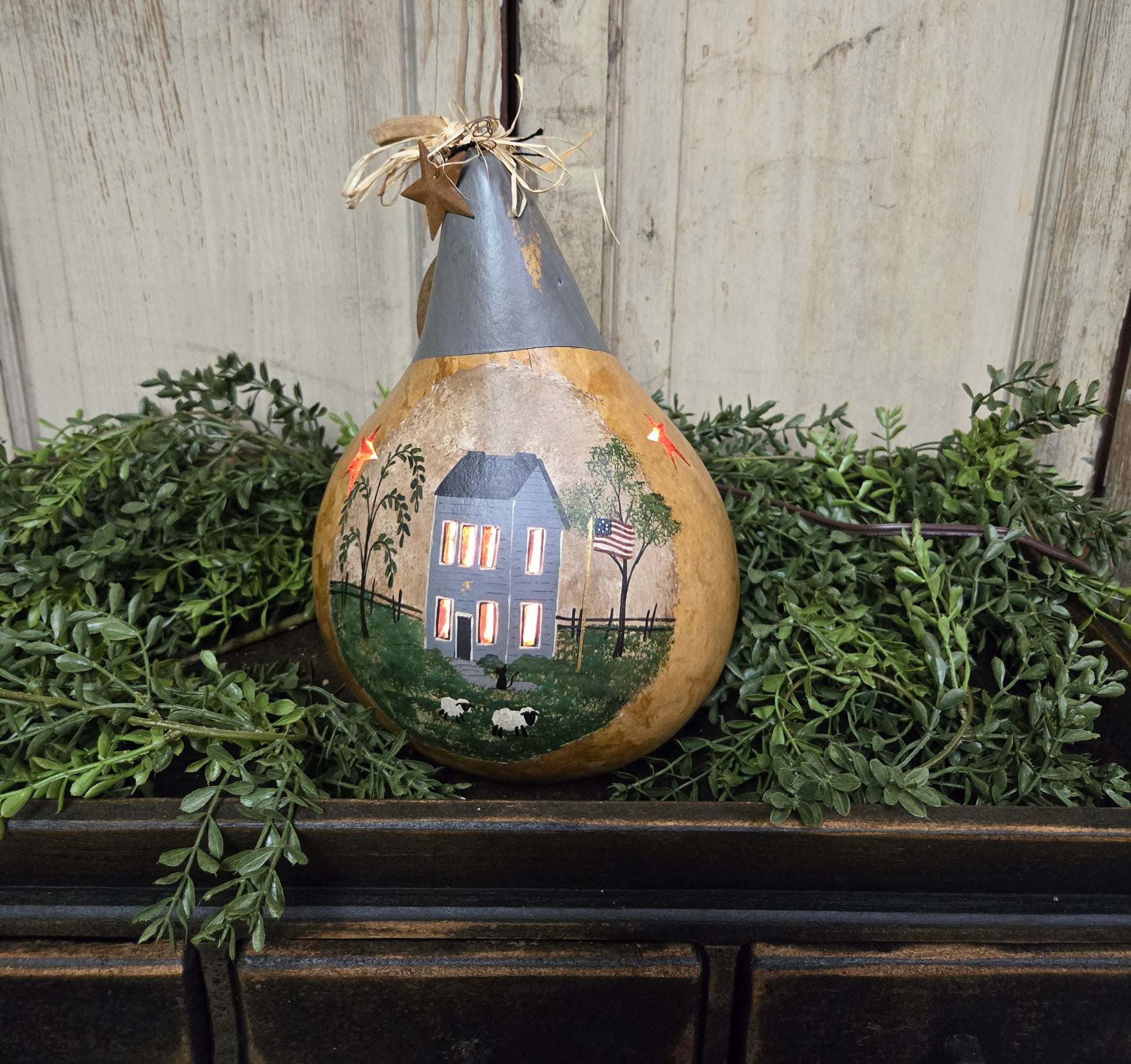 A hand-painted lighted gourd with a rural house scene illuminated from within, placed on a wooden surface with green foliage around it.