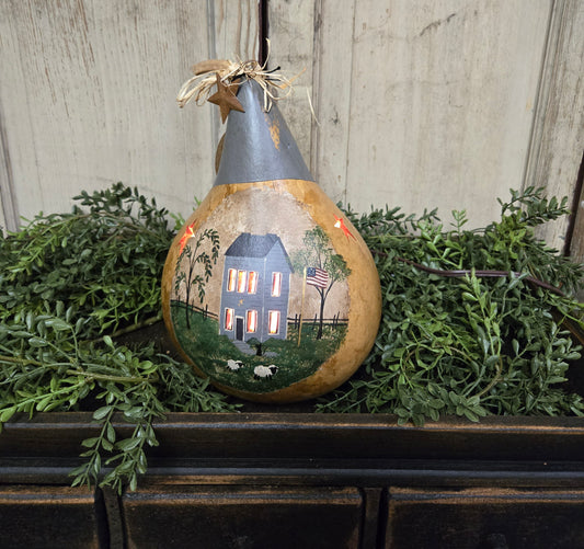 A hand-painted lighted gourd with a rural house scene illuminated from within, placed on a wooden surface with green foliage around it.