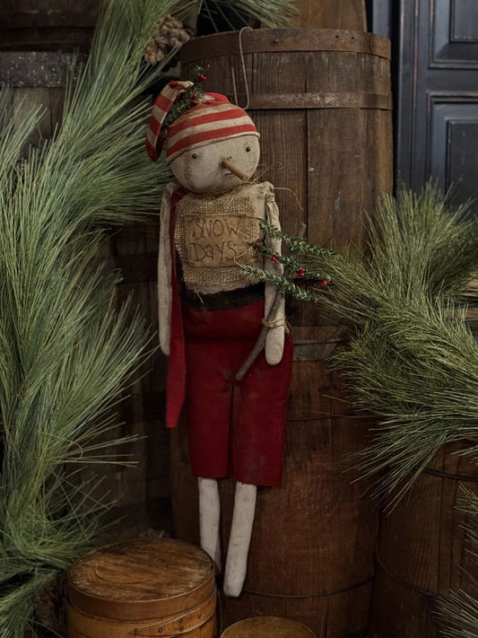 A rustic Christmas rag doll figurine wearing a red and white striped hat and red pants, holding a burlap sign that says 'SNOW DAYS' and a sprig of greenery, posed in front of wooden barrels with pine branches.