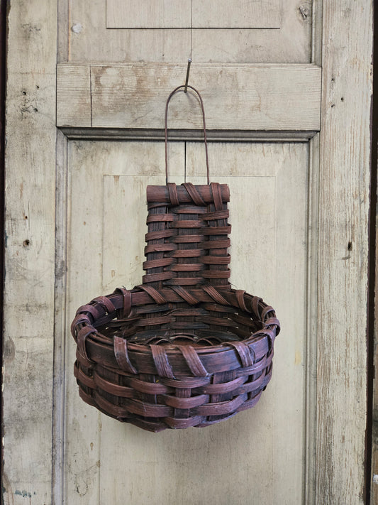 A handmade Primitive Cupboard Basket with a burgundy color, hanging against a rustic wooden background.