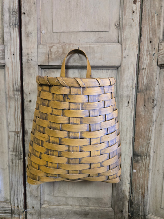 A primitive deep door basket in mustard color, hanging against a wooden background.