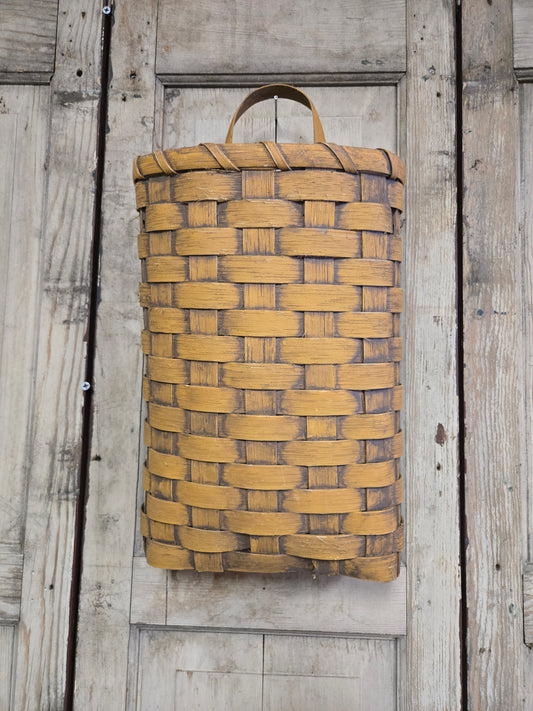 A woven basket hanging on a wooden door, primarily beige in color with brown stripes, likely handcrafted.