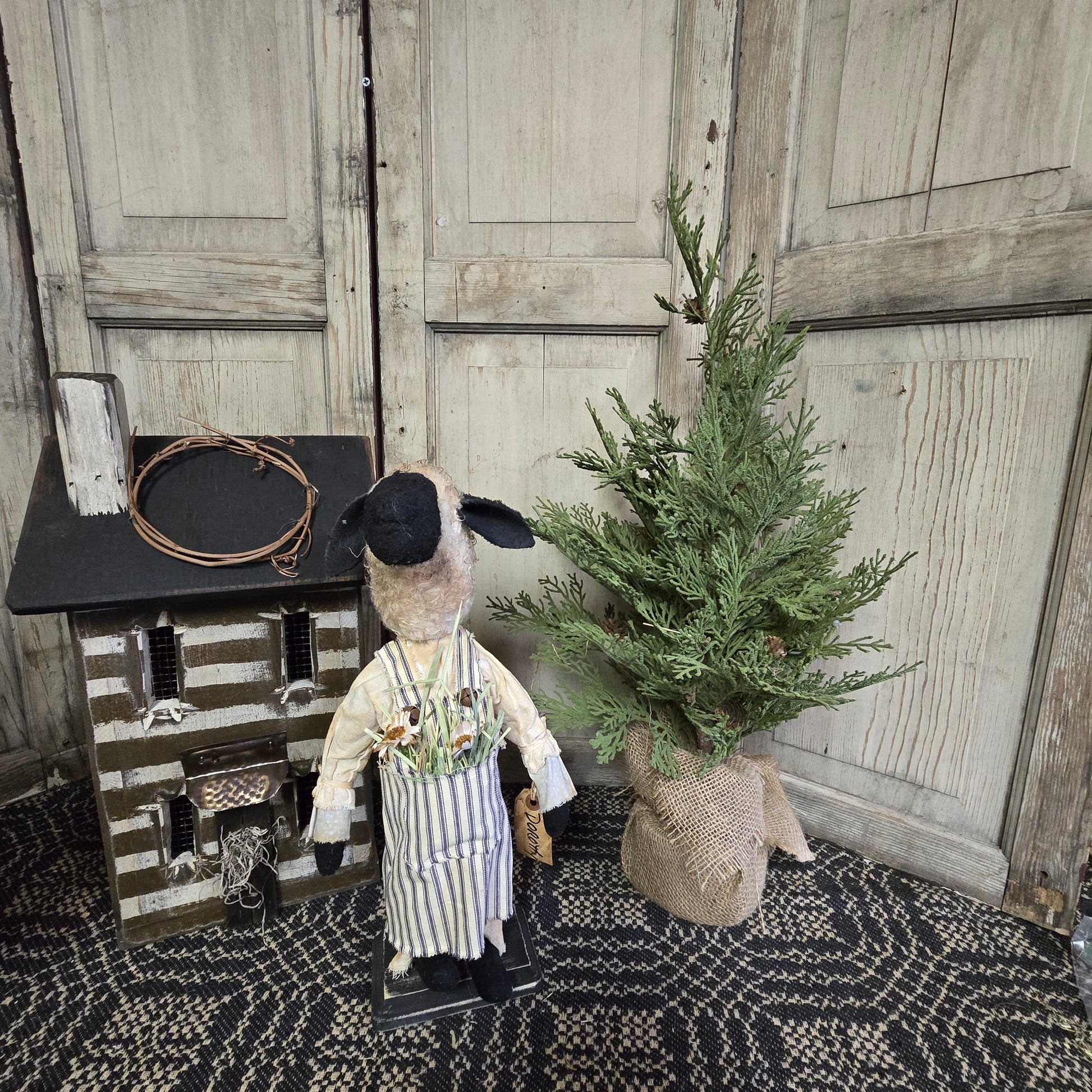 A handcrafted Primitive Dorothy Sheep figurine, wearing a striped outfit and a hat, standing next to a decorative tree.