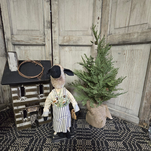 A handcrafted Primitive Dorothy Sheep figurine, wearing a striped outfit and a hat, standing next to a decorative tree.