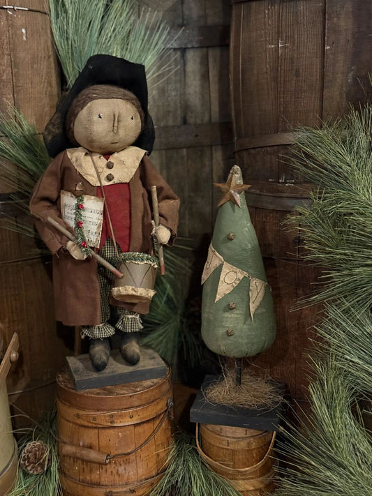 A rustic holiday decorative scene with a cloth doll dressed in brown and red clothing standing on a wooden barrel, next to a small green Christmas tree with a star topper, set against a wooden wall with pine branches.