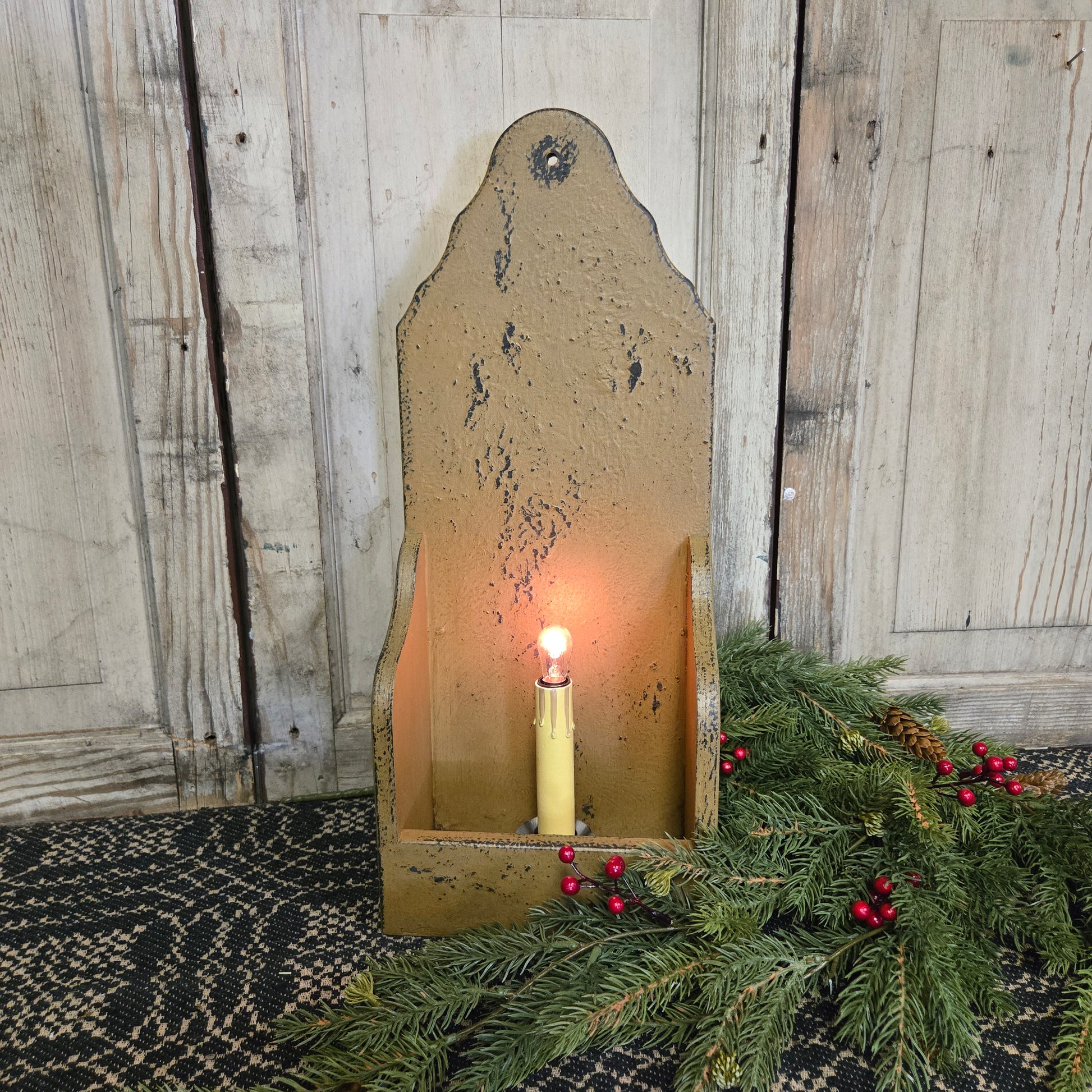 A rustic, mustard-colored wooden candle holder designed for an electric candlestick, displayed with a lit candlestick inside it, surrounded by decorative greenery.