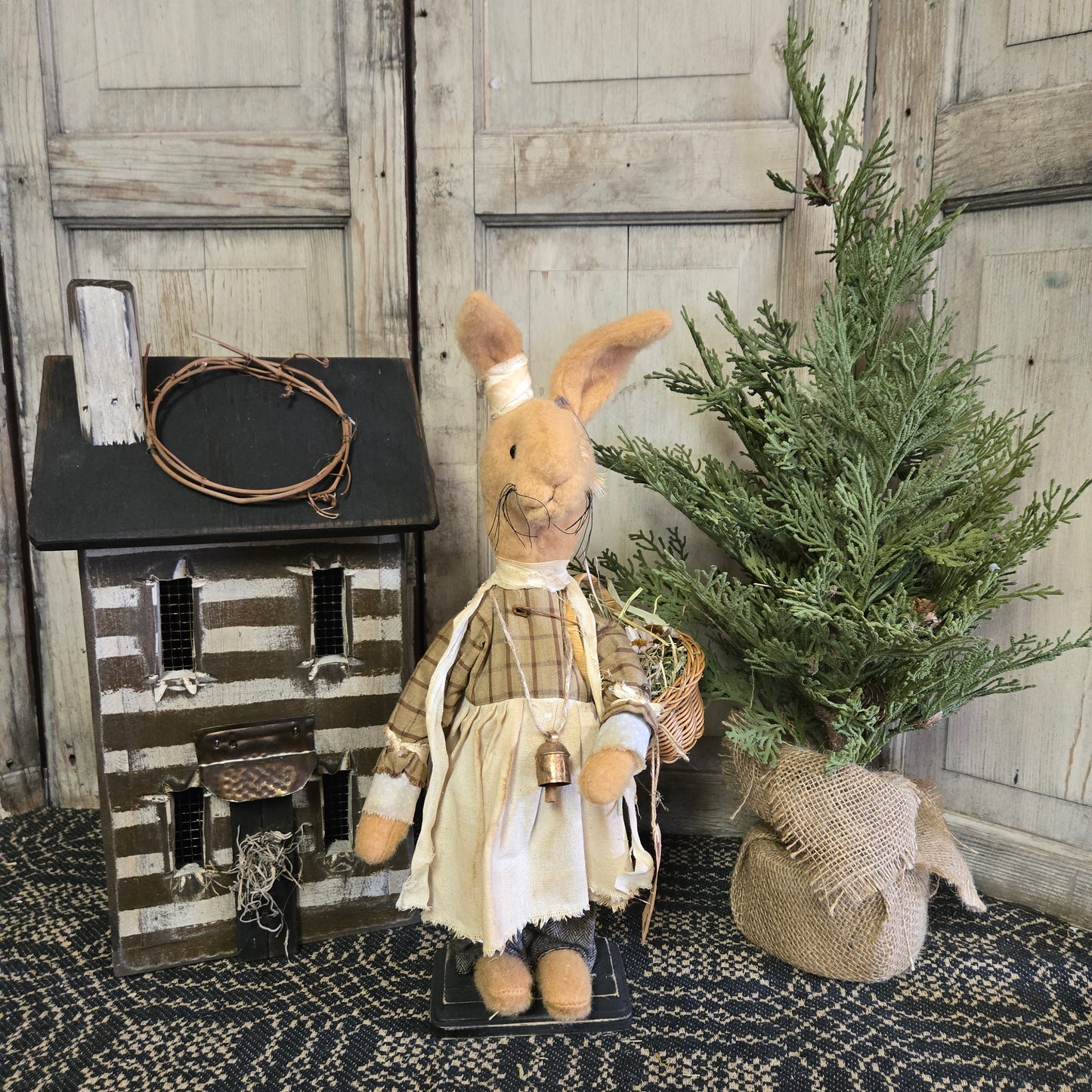 A handcrafted Primitive Emma Rabbit figurine with a basket of flowers, positioned beside a rustic wooden cabinet and a decorative burlap bag.
