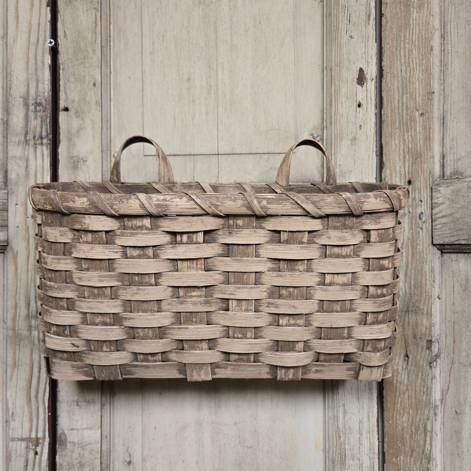 A beige woven basket with handles, hanging against a wooden background.
