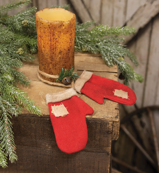 A pair of red fabric mittens with ivory sherpa cuffs and tan linen patches, secured with a thick jute rope, adorned with faux pine and a rusted jingle bell, displayed on a wooden surface with a candle and greenery in the background.