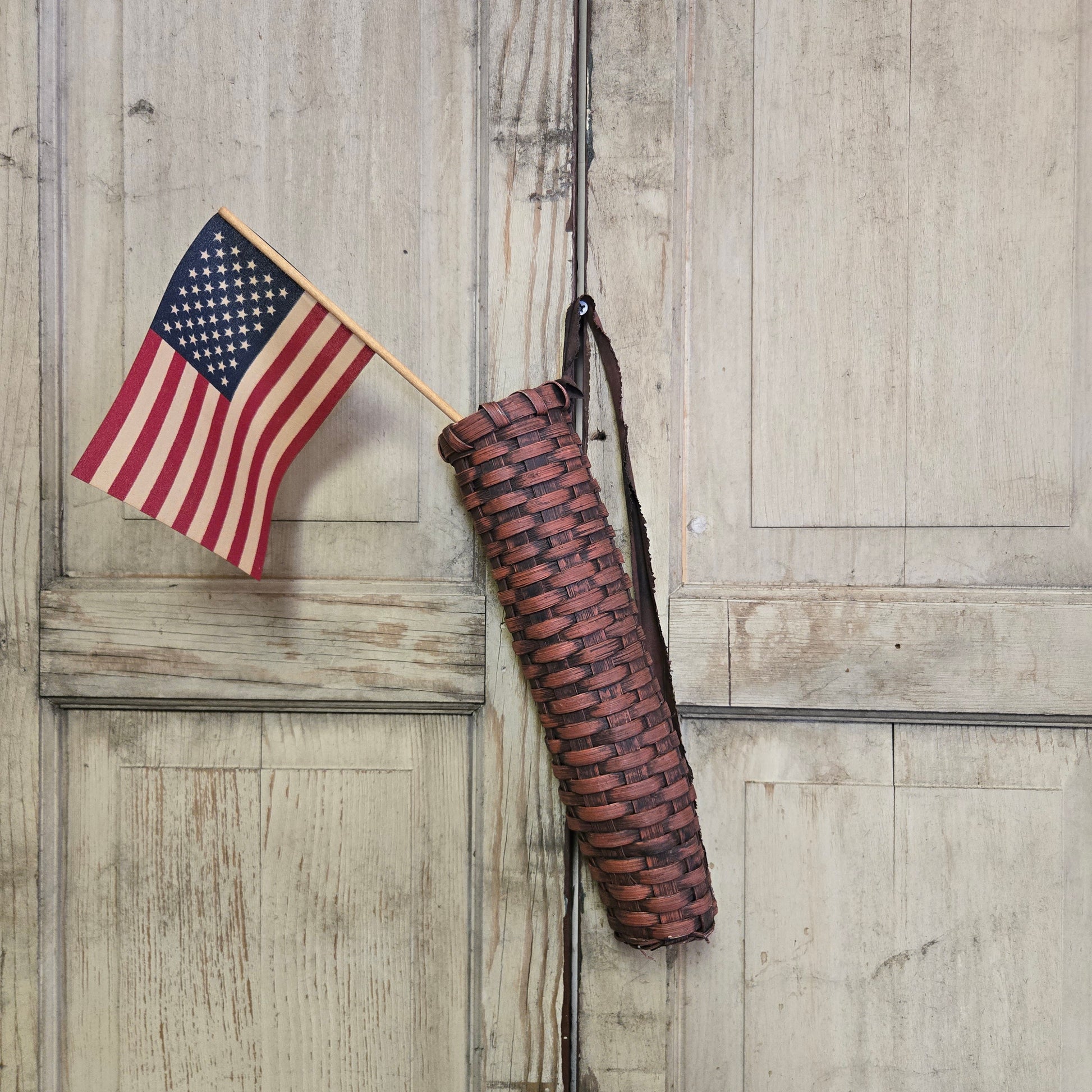 A handwoven burgundy basket with a rustic flag design, displayed against a wooden background. A small American flag is placed next to it.