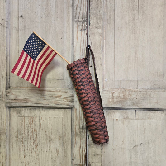 A handwoven burgundy basket with a rustic flag design, displayed against a wooden background. A small American flag is placed next to it.