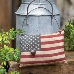 A stuffed fabric pillow ornament designed to resemble an American flag with a vintage look, featuring a brown jingle bell accent.
