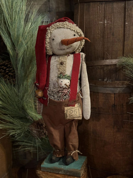A rustic handmade Christmas snowman figurine wearing a red Santa hat and suspenders, brown pants, and a carrot nose, standing on a small teal base in front of a wooden barrel with pine branches.