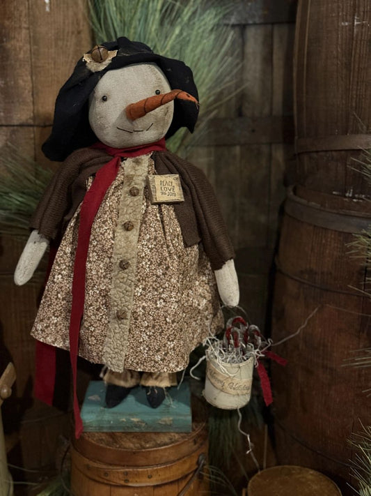 A rustic handmade snowman figurine wearing a black hat, brown coat, and red scarf, holding a small white bucket. The figurine is displayed on a wooden barrel in a themed setting with wood barrels and greenery in the background.