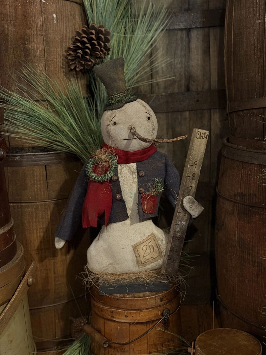 A rustic handmade snowman decoration sitting on a wooden barrel, wearing a dark blue coat, red scarf, and top hat, with pine branches, a pinecone, and a wooden yard sign.
