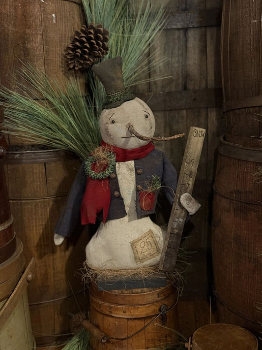 A rustic handmade snowman decoration sitting on a wooden barrel, wearing a dark blue coat, red scarf, and top hat, with pine branches, a pinecone, and a wooden yard sign.