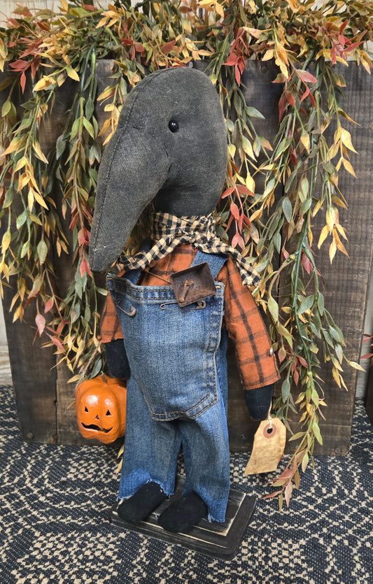 Decorative crow figurine wearing overalls with a pumpkin, surrounded by greenery.