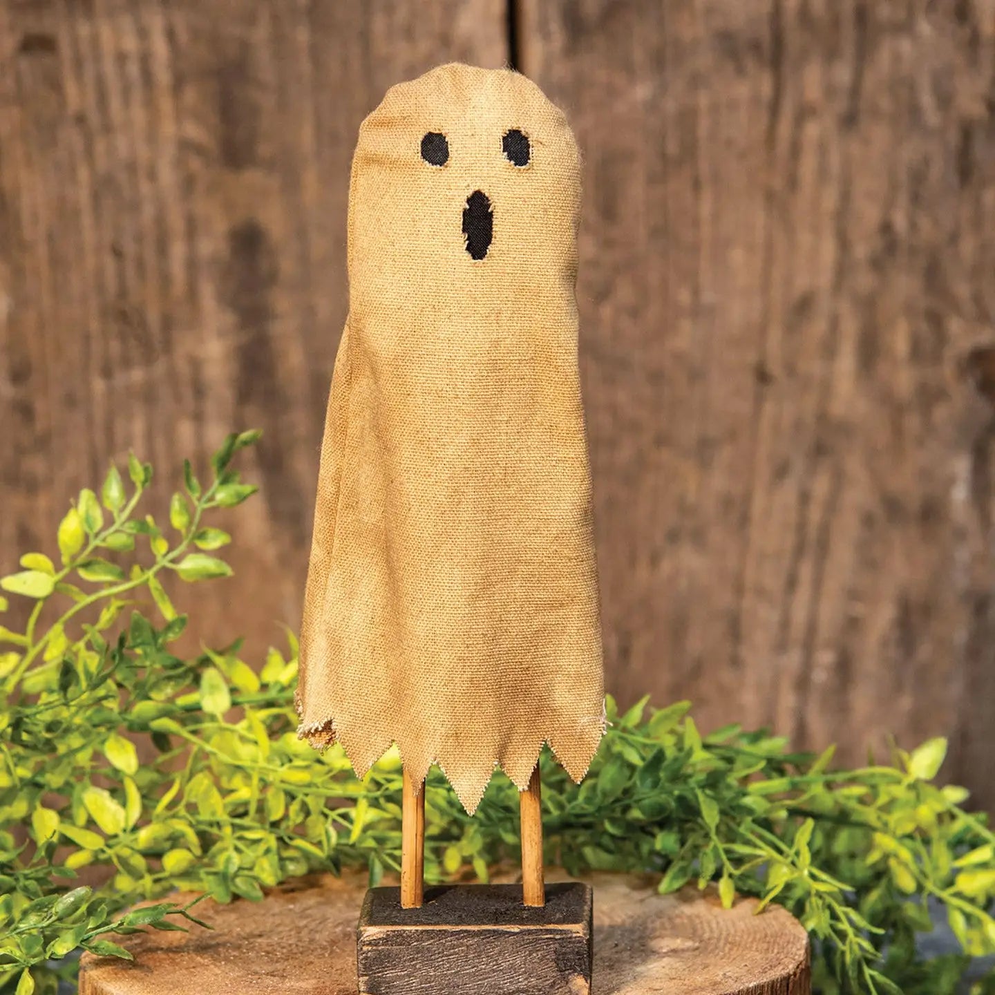 Decorative ghost figure made of burlap with a surprised expression, standing on a wooden base with green foliage in the background.
