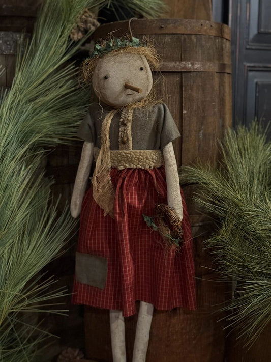 A rustic rag doll with a straw hat and green leafy headpiece, wearing a brown top and red plaid skirt, posed in front of a wooden barrel with pine branches around it.