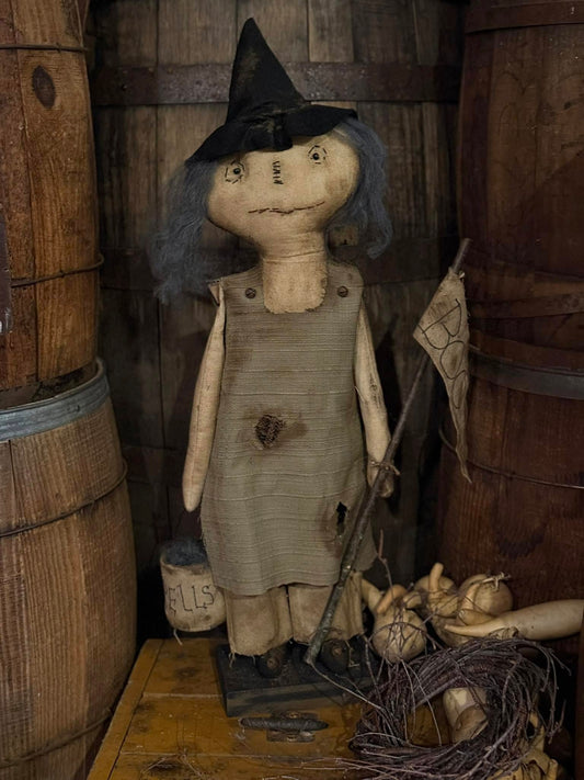 Vintage-style doll with witch outfit and broomstick in a rustic setting with wooden barrels.