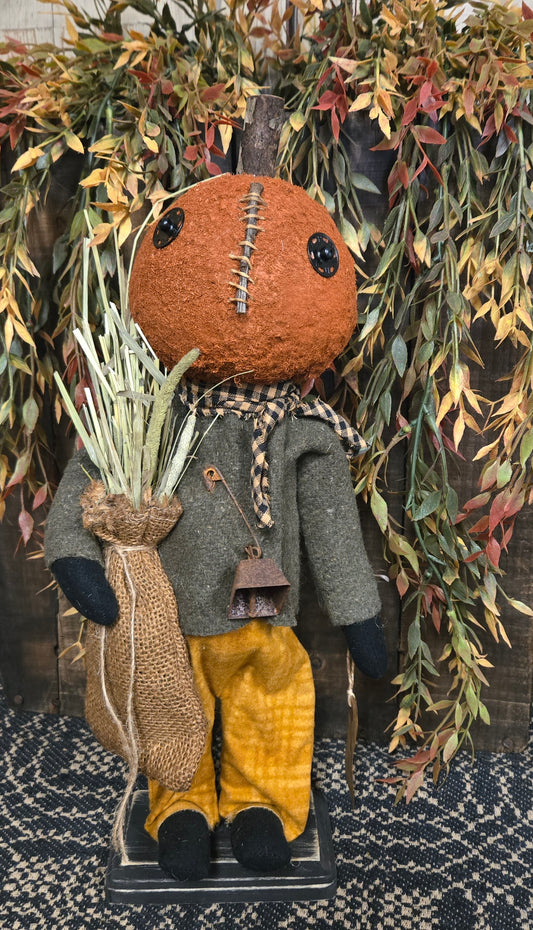 Pumpkin doll with a brown head, gray coat, yellow pants, and black shoes, surrounded by greenery.