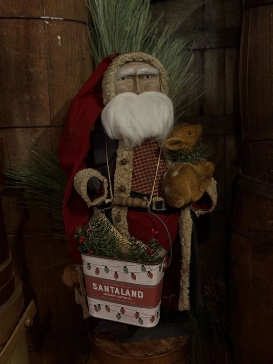 A rustic Santa Claus decorative figure wearing a red suit and hat with a white beard, holding a small reindeer and a white Santa Land tin bucket filled with greenery, set against a dark wooden barrel background.