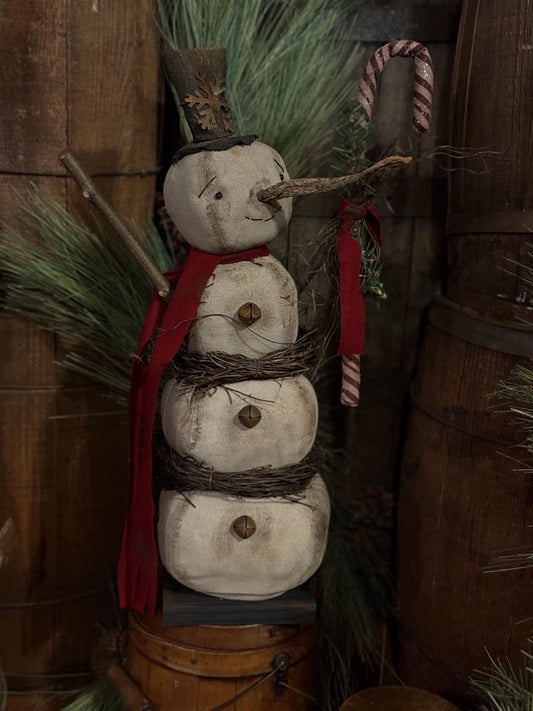 A rustic handmade snowman decoration made from three stacked round natural stones, with a red fabric scarf, twig arms, a small hat, and evergreen branches, displayed against a wooden backdrop with a candy cane prop.