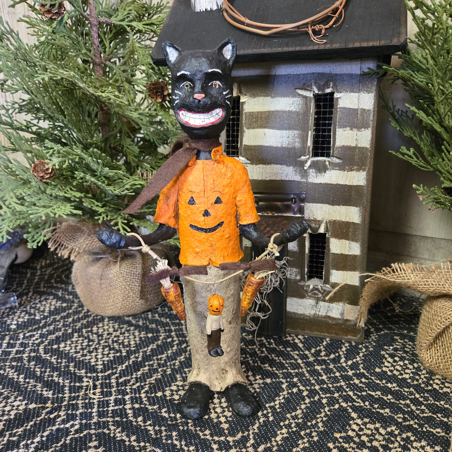 A handcrafted scarecrow with a pumpkin face and black cat features, dressed in an orange shirt and brown pants, standing beside a striped structure.