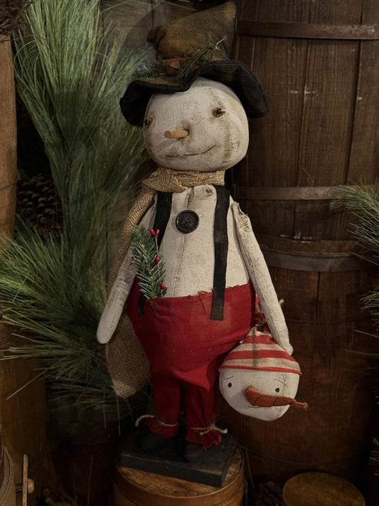 A rustic Christmas decorative doll with a weathered off-white face and hands, wearing a black hat, black suspenders, and red pants, holding a small snowman figure. The doll is styled on a wooden barrel base with pine branches and a wooden barrel in the background.
