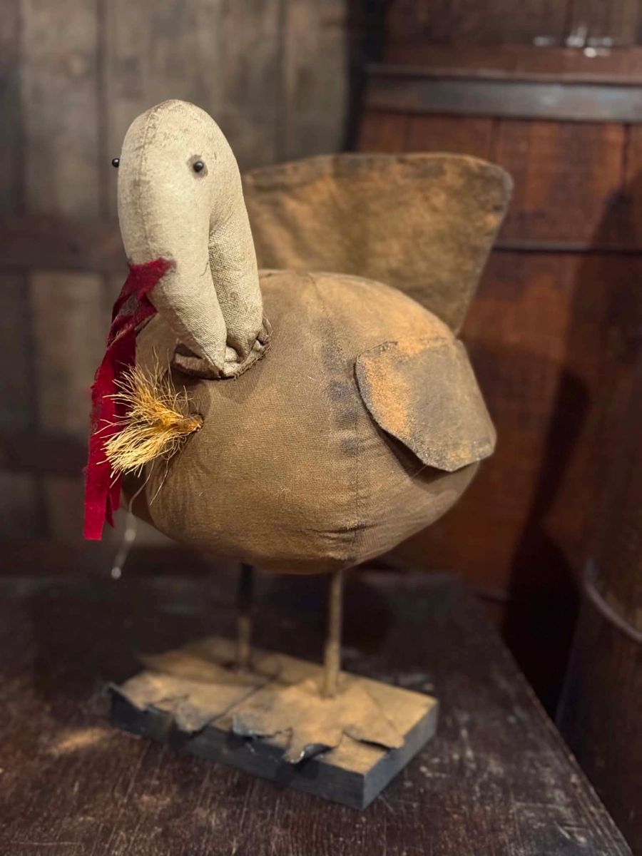 A rustic handmade fabric duck ornament with a tan body, beige head, small black eyes, a red feather detail, and a wooden base, displayed on a dark wooden surface.