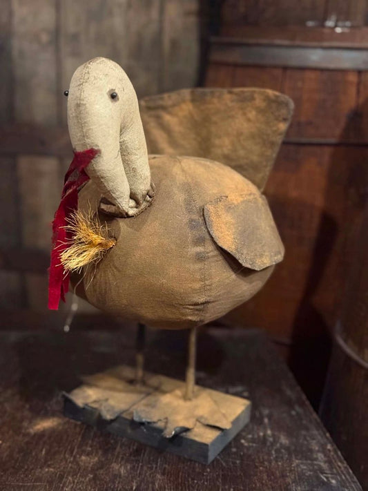 A rustic handmade fabric duck ornament with a tan body, beige head, small black eyes, a red feather detail, and a wooden base, displayed on a dark wooden surface.