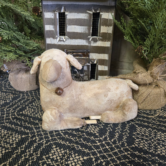 A beige, handcrafted primitive style laying sheep figurine made of wood and fabric, positioned on a textured surface with holiday decorations in the background.