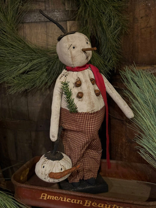 Decorative snowman figure with a red scarf, checkered pants, and a small pumpkin on a wooden surface.