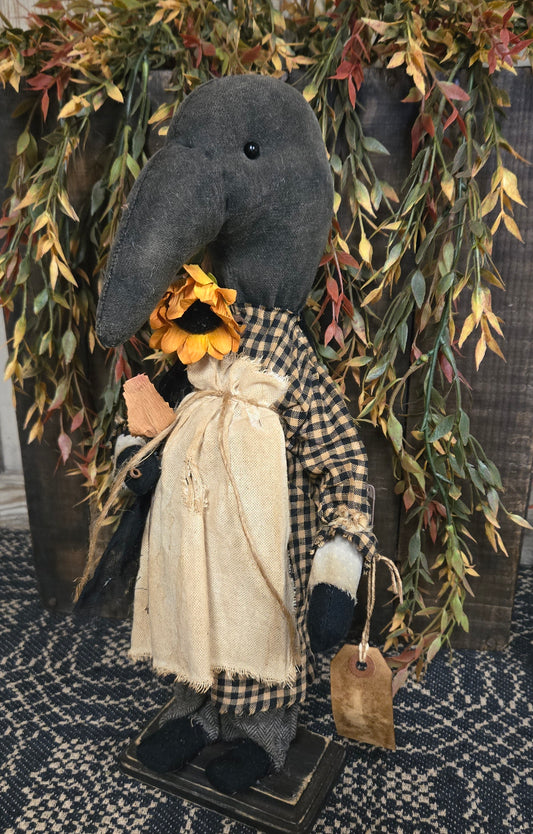 Decorative figure of a crow in a plaid outfit with a sunflower, surrounded by foliage.