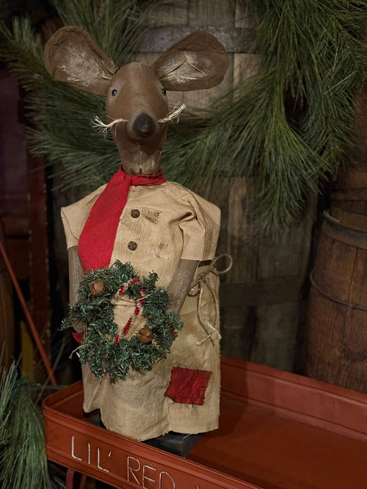 Decorative mouse figure in a coat with a wreath, standing on a wooden surface with greenery in the background.