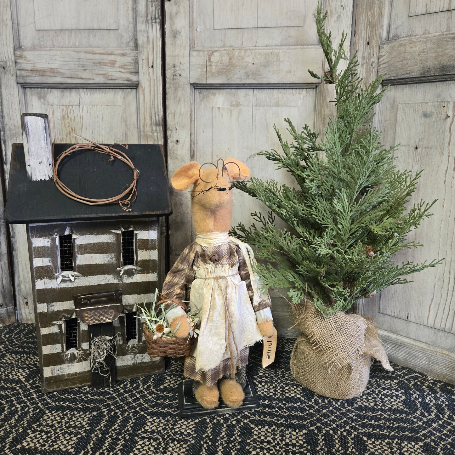 A figurine of a mouse holding a basket of flowers, wearing a plaid shirt and a white apron, placed next to a pine tree decoration.