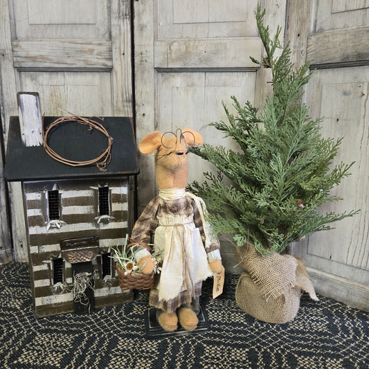 A figurine of a mouse holding a basket of flowers, wearing a plaid shirt and a white apron, placed next to a pine tree decoration.