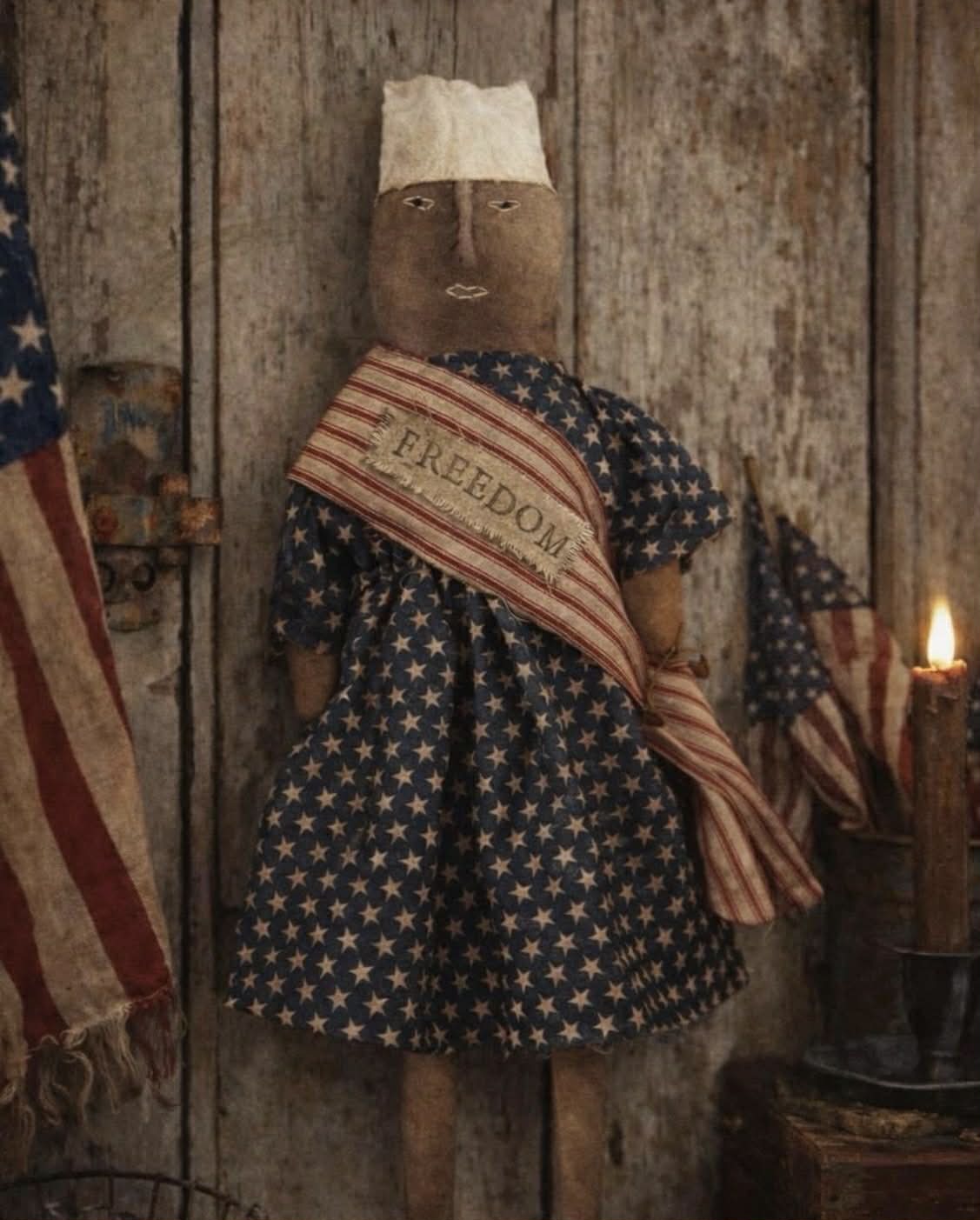 Vintage-style doll with 'Freedom' scarf in a rustic setting with American flags.