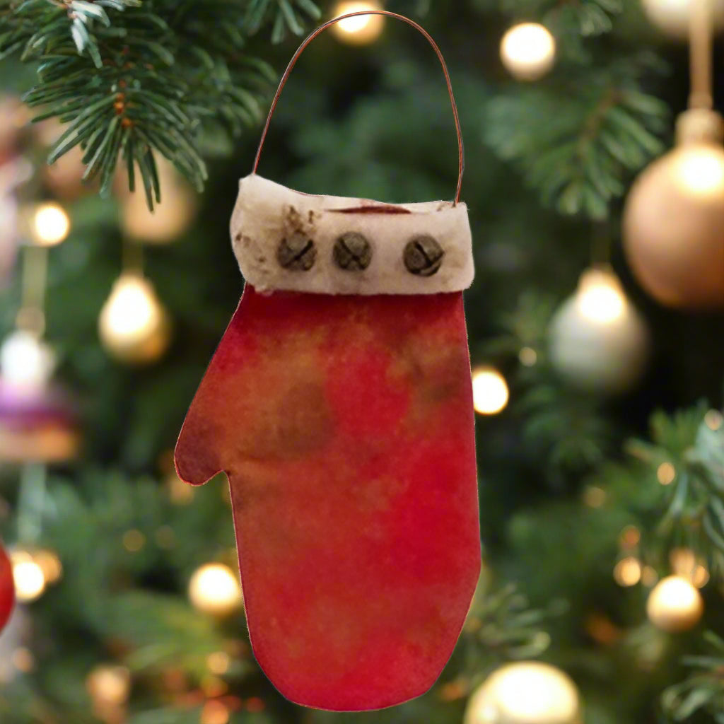 A red fabric mitten with a white cuff and metal bells, with a distressed look and cinnamon dusting for texture, designed as a pocket ornament with a metal wire hanger.