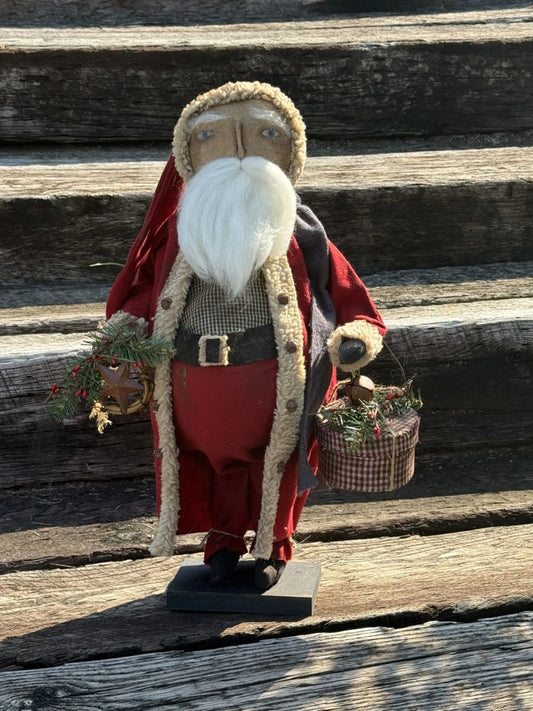 A rustic wooden Santa Claus figurine with a long white beard, red coat, and beige fur trim, standing on a weathered wooden surface and holding small holiday baskets.
