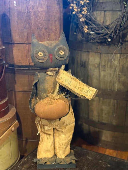 A rustic owl figurine holding a small pumpkin and a handwritten sign that reads 'Give Thanks,' displayed in a vintage-style setting with wooden barrels in the background.