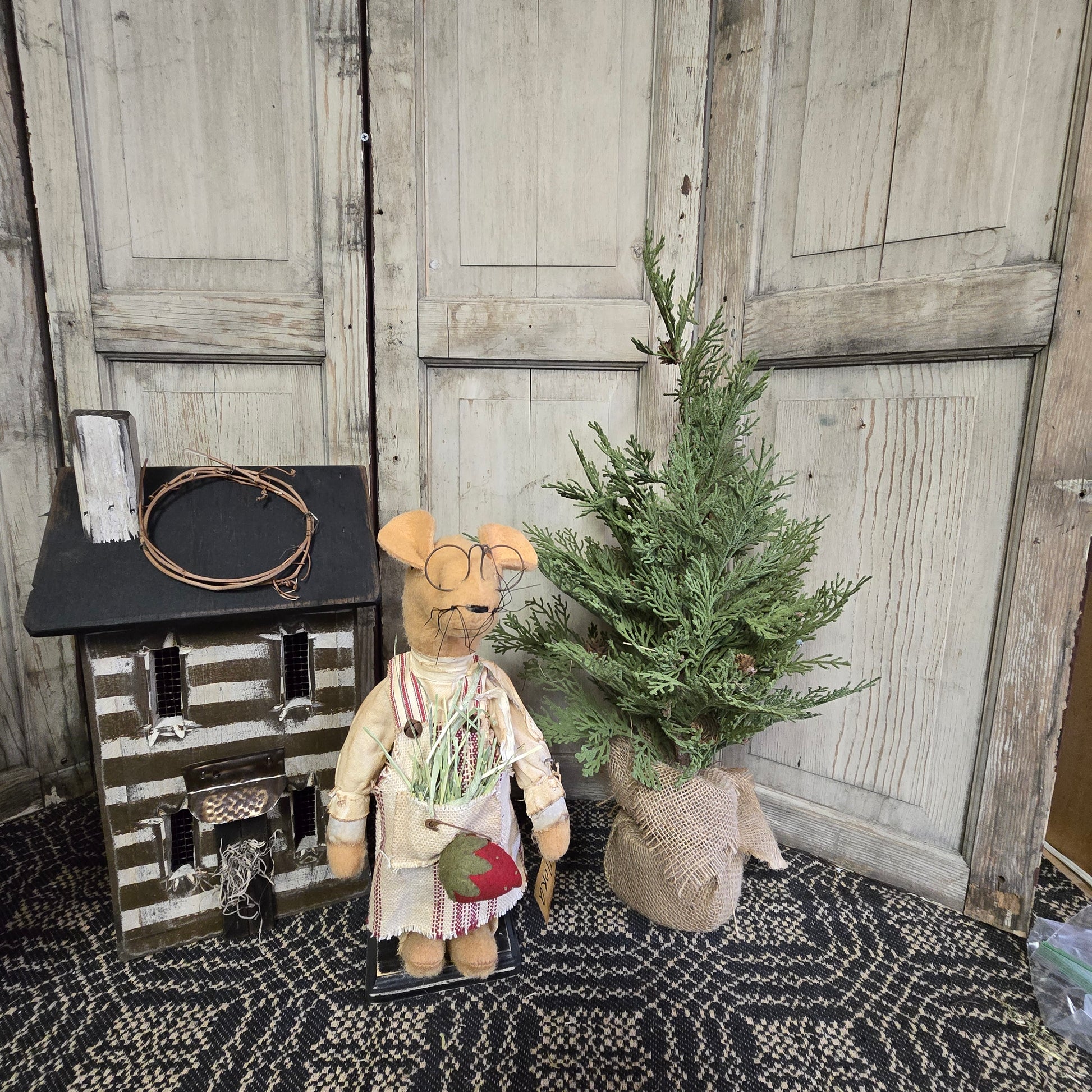 A decorative figurine of a mouse wearing a shirt and holding a strawberry, named Primitive Nora Mouse, displayed beside a miniature wooden door and a small artificial greenery.