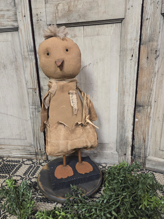 A primitive style sculpture of a chicken with a realistic design, standing against a wooden backdrop with greenery on the ground.