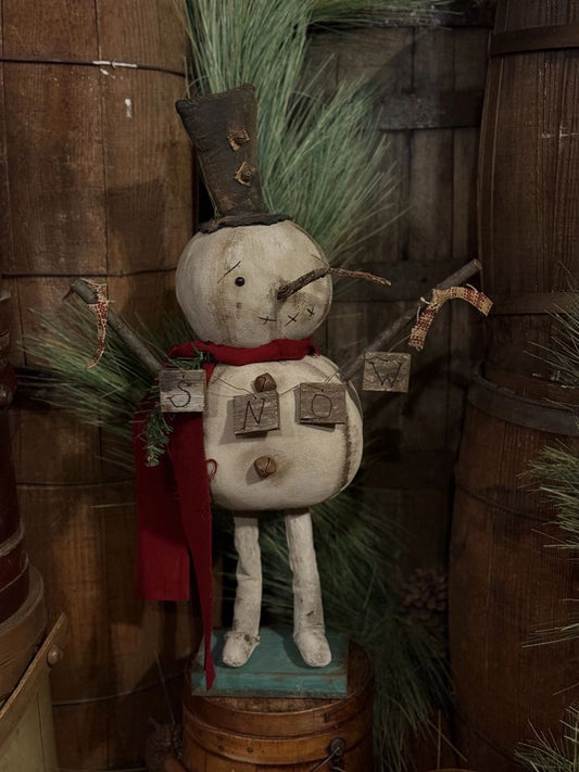 A rustic wooden snowman figurine with a painted off-white body, red scarf, green pine branches for hair, and small hanging tags, displayed against a backdrop of wooden barrels and greenery.
