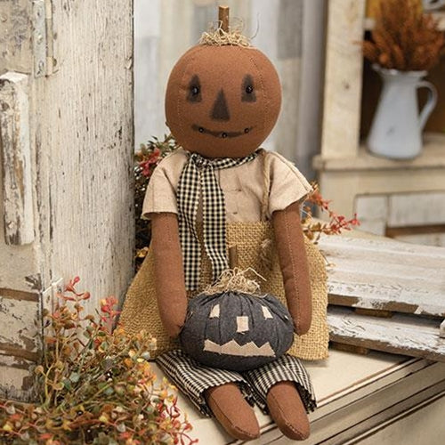 A plush doll resembling a girl with a pumpkin head and a jack-o-lantern face, dressed in a linen and burlap outfit, holding a plush gray jack-o-lantern pumpkin. The doll is seated against a backdrop of a rustic wooden setting with flowers.