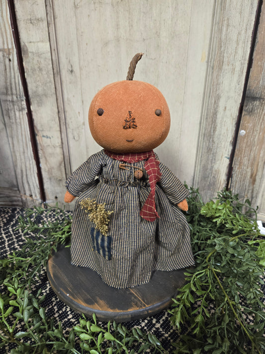 Primitive pumpkin girl in navy dress with greenery in background.